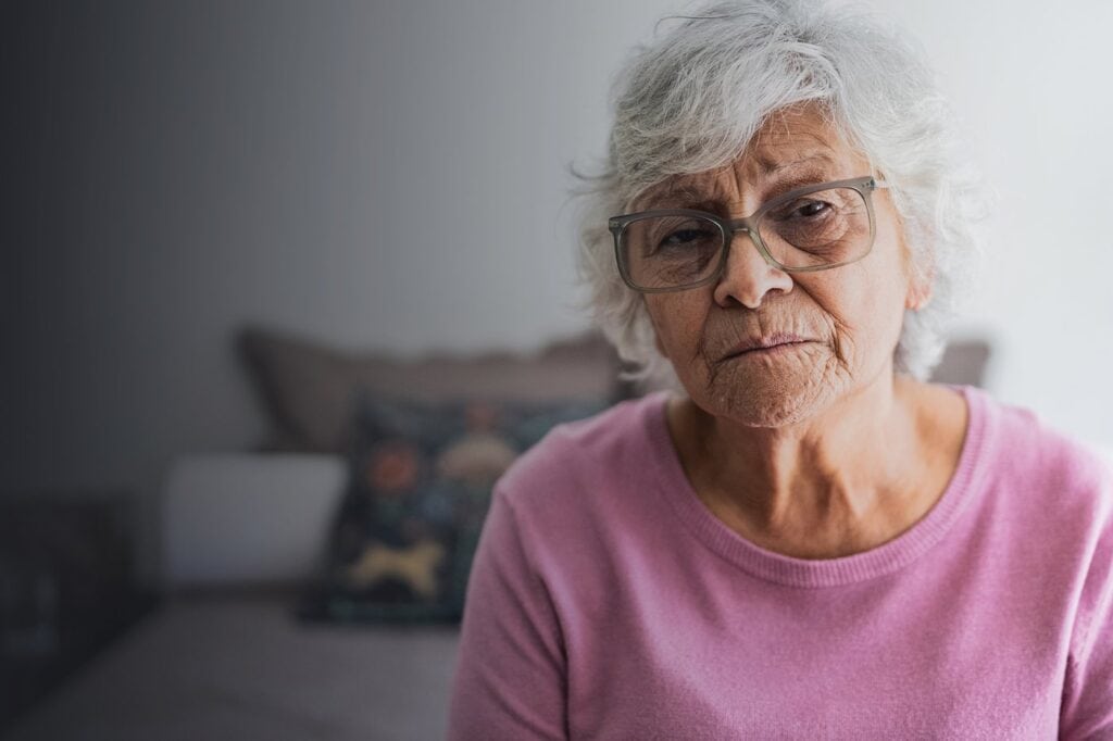An older woman with grey hair and glasses, sitting in her living room